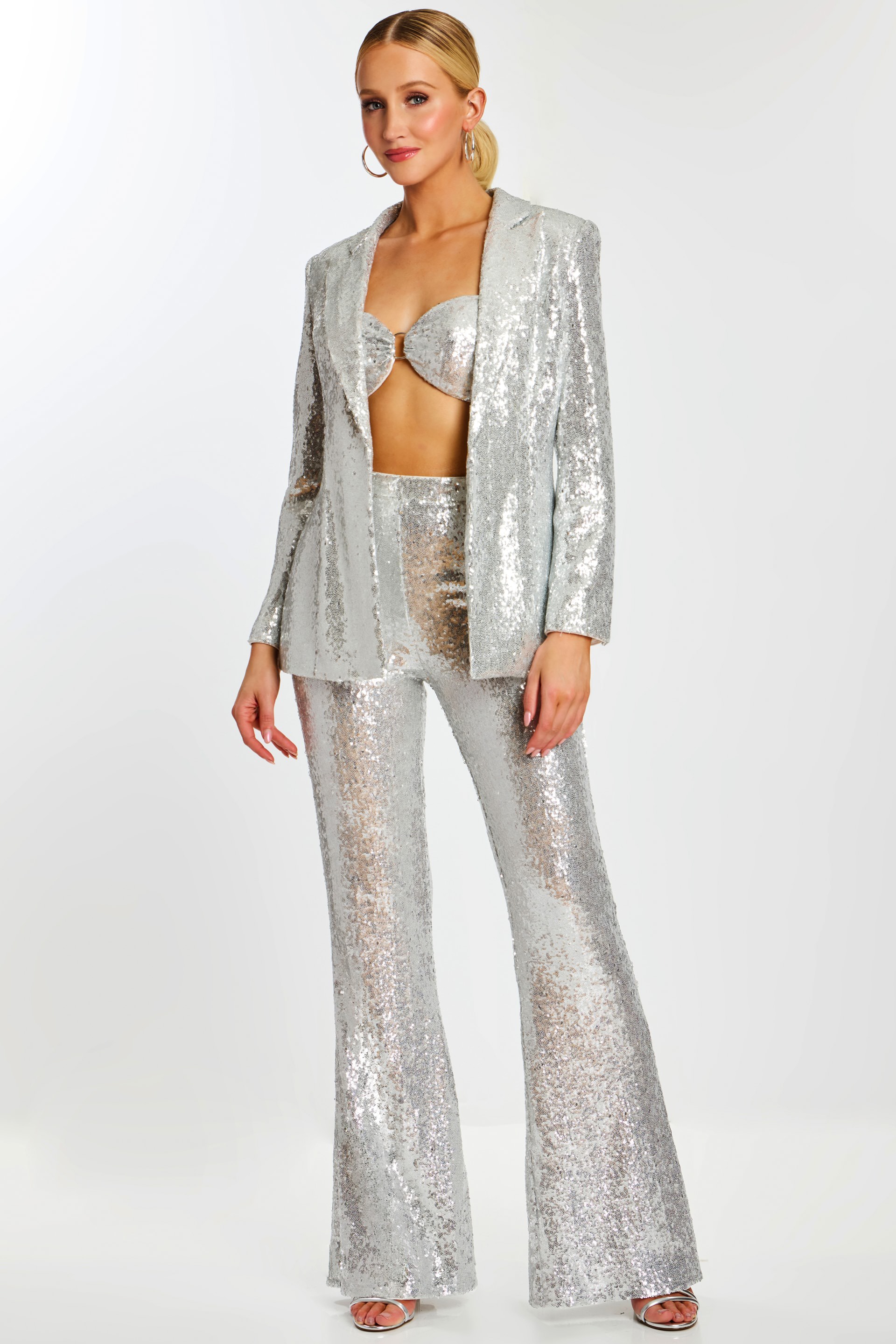 Model wearing silver sequin pantsuit with flared trousers, bralette top, and open blazer.