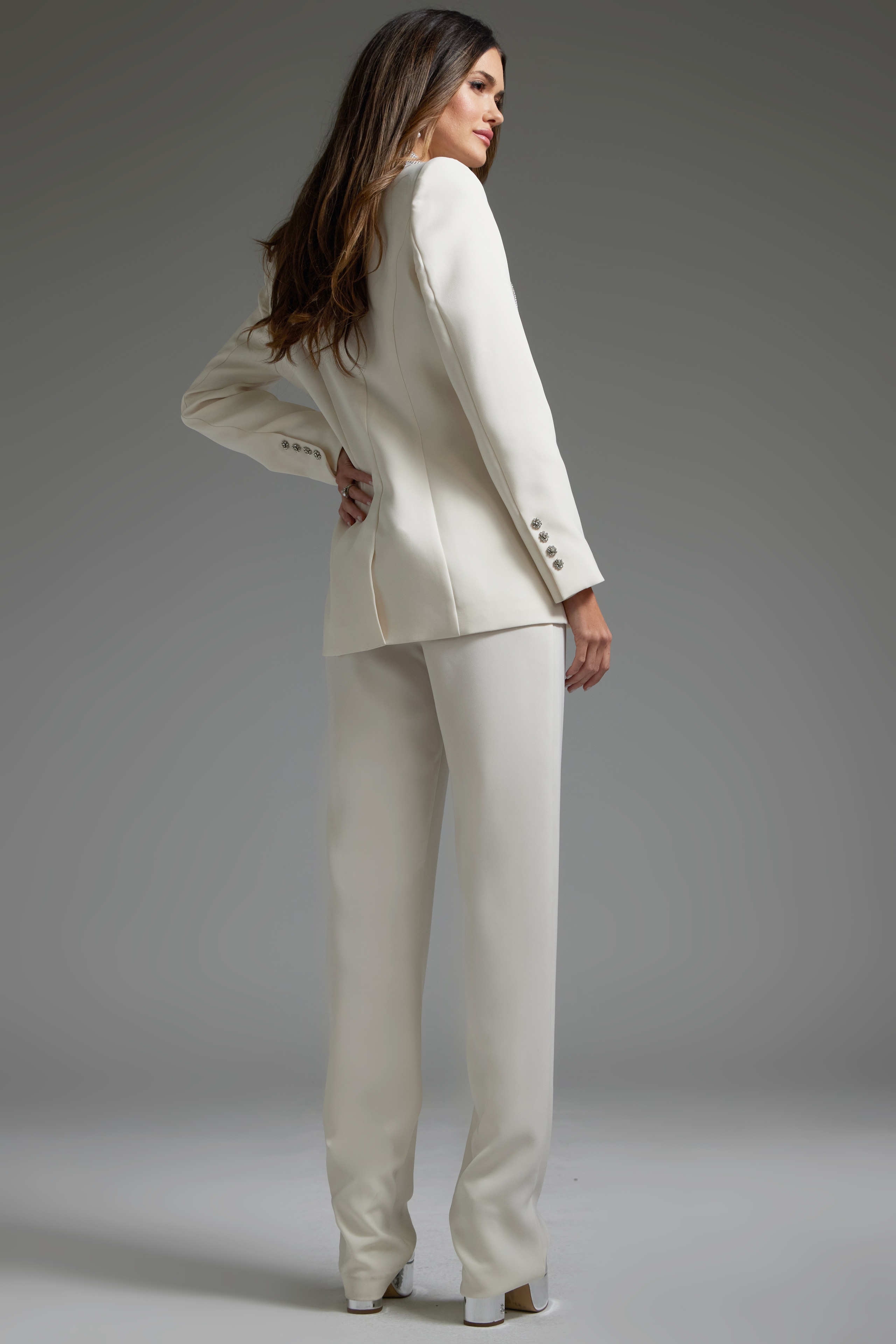 Model wearing Jovani 39726 ivory pant suit with silver embellishments, back view.