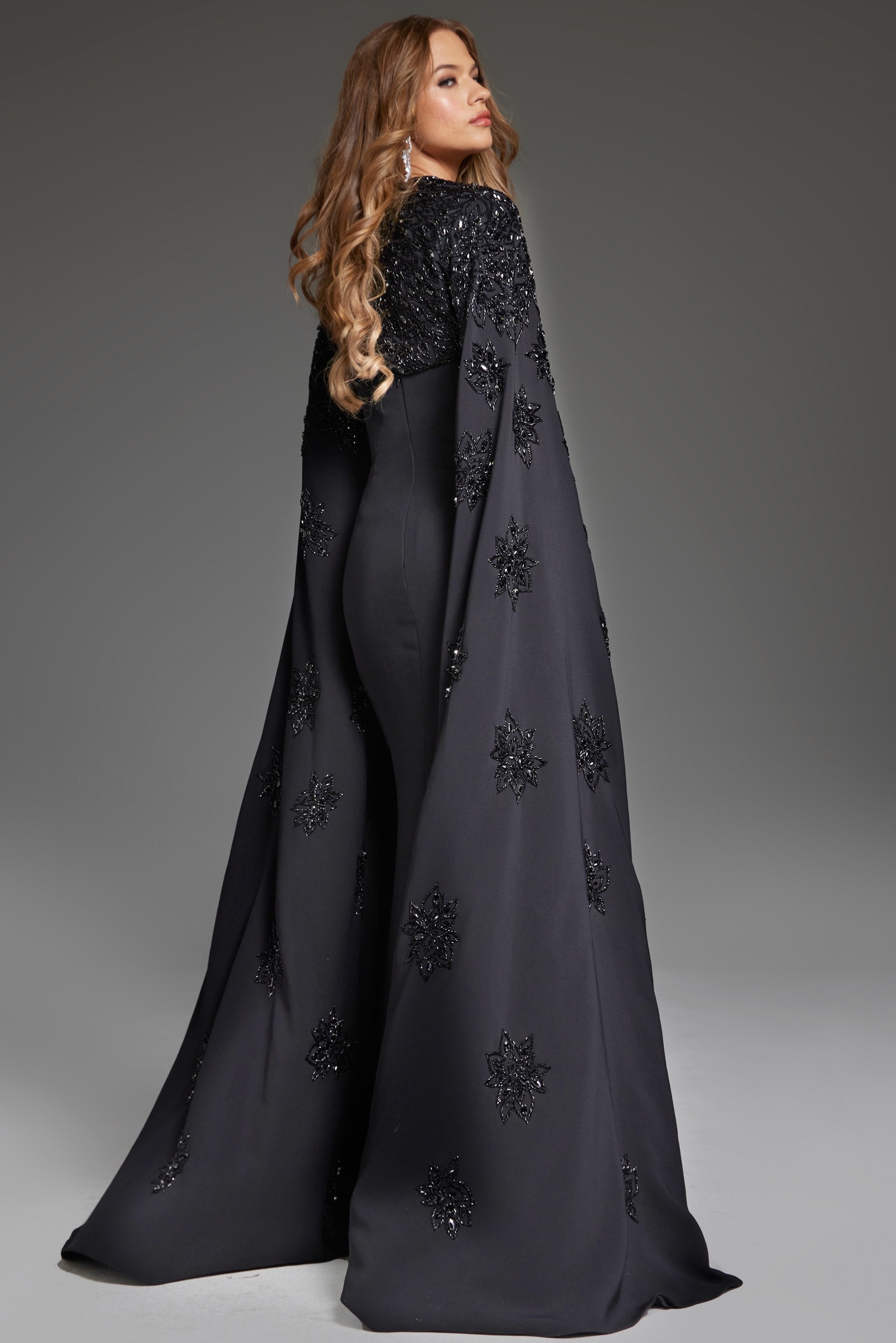back view of model wearing Jovani 39957 black gown highlighting the dramatic cape