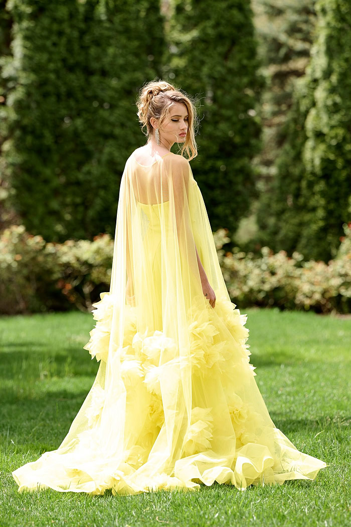 Model wearing Jovani 40092 yellow gown with flowing tulle and floral details, angled back view