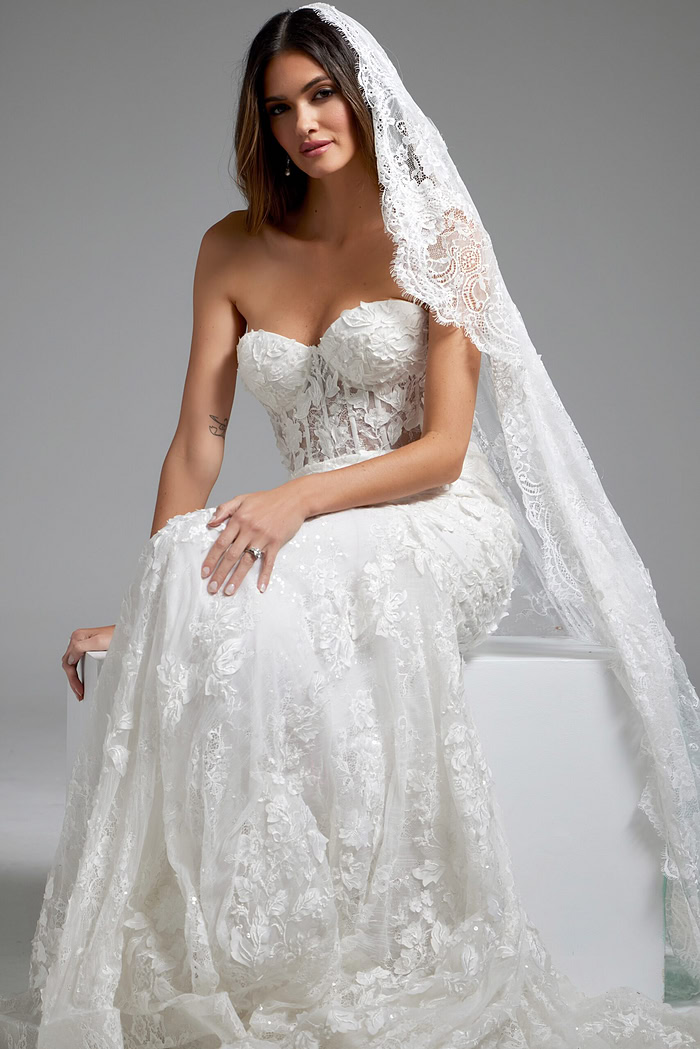 Model wearing Jovani JB07583 white gown sitting, showcasing strapless neckline and lace embroidery.